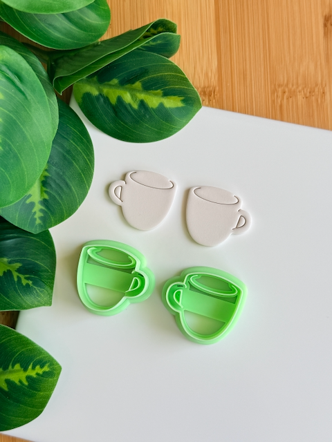 Coffee mug polymer clay cutters and white clay impressions on a white surface next to green leaves.