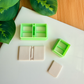Two light green 3D-printed polymer clay cutters on a white surface. One cutter features an integrated design for a classic book spine with blank covers, while the other is a simple rectangular cutter. Below the tools are two light grey clay samples: one featuring the embossed book spine and a smaller, plain rectangular piece. Green leaves and a wooden surface are visible in the background.
