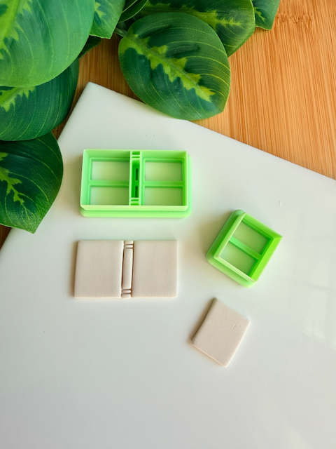 Two light green 3D-printed polymer clay cutters on a white surface. One cutter features an integrated design for a classic book spine with blank covers, while the other is a simple rectangular cutter. Below the tools are two light grey clay samples: one featuring the embossed book spine and a smaller, plain rectangular piece. Green leaves and a wooden surface are visible in the background.