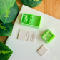 Two light green 3D-printed polymer clay cutters on a white surface. One cutter includes an integrated design for a book spine and a melting candle with a flame on the cover. Next to the tools are two light grey clay samples: one with the embossed candle book impression and one plain rectangular piece. Green leaves and a wooden surface are visible in the background.