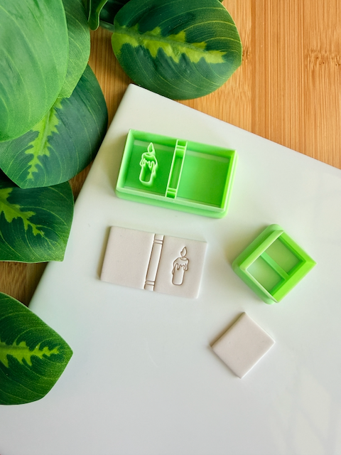Two light green 3D-printed polymer clay cutters on a white surface. One cutter includes an integrated design for a book spine and a melting candle with a flame on the cover. Next to the tools are two light grey clay samples: one with the embossed candle book impression and one plain rectangular piece. Green leaves and a wooden surface are visible in the background.