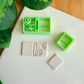 Two light green 3D-printed polymer clay cutters on a white surface. One cutter features an integrated design for a book spine and two butterflies on the cover. Next to them are two light grey clay samples: one with the embossed butterfly book design and a smaller, plain rectangular piece. Green leaves and a wooden surface are visible in the background.