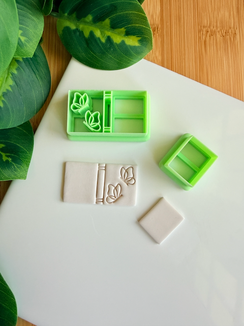 Two light green 3D-printed polymer clay cutters on a white surface. One cutter features an integrated design for a book spine and two butterflies on the cover. Next to them are two light grey clay samples: one with the embossed butterfly book design and a smaller, plain rectangular piece. Green leaves and a wooden surface are visible in the background.