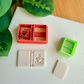 A reddish-orange 3D-printed polymer clay cutter featuring an integrated design for a book spine and a steaming coffee cup on the cover. Below it is a light grey clay sample with the embossed coffee book design. A small light green rectangular cutter and a plain rectangular clay piece are also shown on the white surface, framed by green leaves and a wooden background.