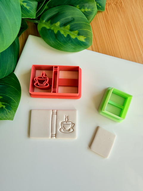 A reddish-orange 3D-printed polymer clay cutter featuring an integrated design for a book spine and a steaming coffee cup on the cover. Below it is a light grey clay sample with the embossed coffee book design. A small light green rectangular cutter and a plain rectangular clay piece are also shown on the white surface, framed by green leaves and a wooden background.