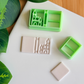 Two light green 3D-printed polymer clay cutters on a white surface. One cutter includes an integrated stamp featuring a book spine and a detailed castle silhouette. Next to the tools are two samples of light grey clay: one with the embossed castle book impression and one plain rectangular piece. Green leaves are visible in the top left corner against a wooden background.