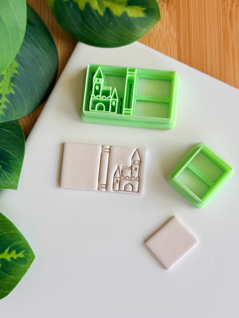 Two light green 3D-printed polymer clay cutters on a white surface. One cutter includes an integrated stamp featuring a book spine and a detailed castle silhouette. Next to the tools are two samples of light grey clay: one with the embossed castle book impression and one plain rectangular piece. Green leaves are visible in the top left corner against a wooden background.