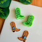 A mirrored pair of light green 3D-printed cowboy boot clay cutters with an intricate leaf and vine "embroidery" pattern. Below the tools are two finished brown clay boot samples featuring the embossed design and decorated with small handmade white flowers at the ankle. The scene is framed by green leaves on a white surface with a wooden background.