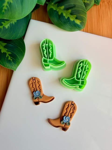 A mirrored pair of light green 3D-printed cowboy boot clay cutters with an intricate leaf and vine "embroidery" pattern. Below the tools are two finished brown clay boot samples featuring the embossed design and decorated with small handmade white flowers at the ankle. The scene is framed by green leaves on a white surface with a wooden background.