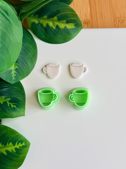 Coffee mug polymer clay cutters and white clay impressions on a white surface next to green leaves.