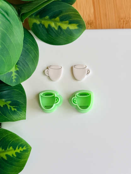 Coffee mug polymer clay cutters and white clay impressions on a white surface next to green leaves.