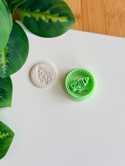 Spiral seashell clay cutter and its white clay impression on a white surface next to green leaves.