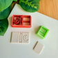A reddish-orange 3D-printed polymer clay cutter with an integrated design for a book spine and a crescent moon on the cover. Below it is a light grey clay sample with the embossed crescent moon book design. A small light green rectangular cutter and a plain rectangular clay piece are also shown on the white surface, with green leaves and a wooden surface in the background.