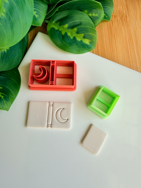 A reddish-orange 3D-printed polymer clay cutter with an integrated design for a book spine and a crescent moon on the cover. Below it is a light grey clay sample with the embossed crescent moon book design. A small light green rectangular cutter and a plain rectangular clay piece are also shown on the white surface, with green leaves and a wooden surface in the background.