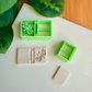 Two light green 3D-printed polymer clay cutters on a white surface. One cutter features an integrated design for a book spine and a detailed dragonfly on the cover. Next to the tools are two light grey clay samples: one with the embossed dragonfly book impression and one plain rectangular piece. Green leaves and a wooden bamboo surface are visible in the background.