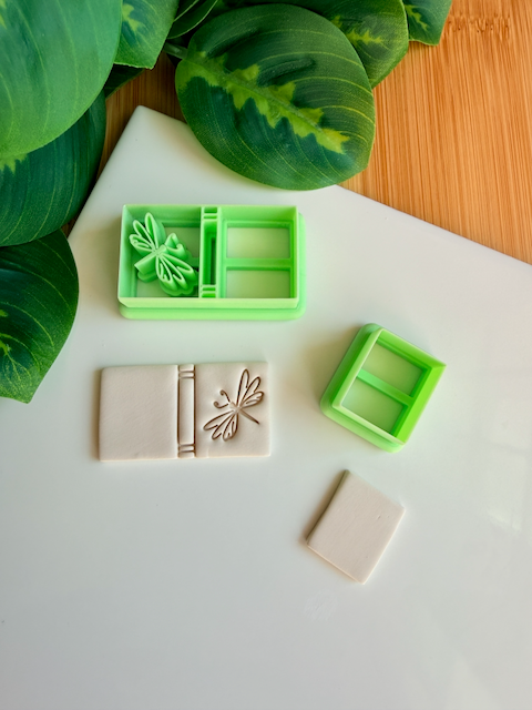 Two light green 3D-printed polymer clay cutters on a white surface. One cutter features an integrated design for a book spine and a detailed dragonfly on the cover. Next to the tools are two light grey clay samples: one with the embossed dragonfly book impression and one plain rectangular piece. Green leaves and a wooden bamboo surface are visible in the background.