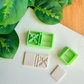 Two light green 3D-printed polymer clay cutters on a white surface. One cutter features an integrated design for a book spine and a bold crossed swords motif on the cover. Below the tools are two light grey clay samples: one with the embossed adventure book design and a smaller, plain rectangular piece. Green leaves and a wooden surface are visible in the background.