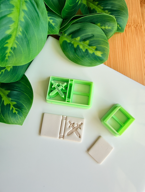 Two light green 3D-printed polymer clay cutters on a white surface. One cutter features an integrated design for a book spine and a bold crossed swords motif on the cover. Below the tools are two light grey clay samples: one with the embossed adventure book design and a smaller, plain rectangular piece. Green leaves and a wooden surface are visible in the background.