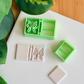 A top-down view of a mint green, multi-piece 3D-printed clay cutter set featuring a "butterfly journal" or "open book" theme. The main rectangular cutter includes a detailed butterfly emblem and a banded spine, shown alongside a white polymer clay piece capturing the crisp, embossed butterfly and book details. A smaller square cutter and a plain clay piece are also part of the arrangement on a clean white surface with green leaves and a wooden background in the corners.