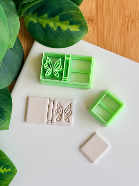 A top-down view of a mint green, multi-piece 3D-printed clay cutter set featuring a "butterfly journal" or "open book" theme. The main rectangular cutter includes a detailed butterfly emblem and a banded spine, shown alongside a white polymer clay piece capturing the crisp, embossed butterfly and book details. A smaller square cutter and a plain clay piece are also part of the arrangement on a clean white surface with green leaves and a wooden background in the corners.