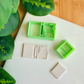 Two light green 3D-printed polymer clay cutters on a white surface. One cutter features an integrated design for a book spine and elegant filigree scrollwork in the corners of the cover. Next to them are two light grey clay samples: one with the embossed ornate book design and a smaller, plain rectangular piece. Green leaves and a wooden surface are visible in the background.