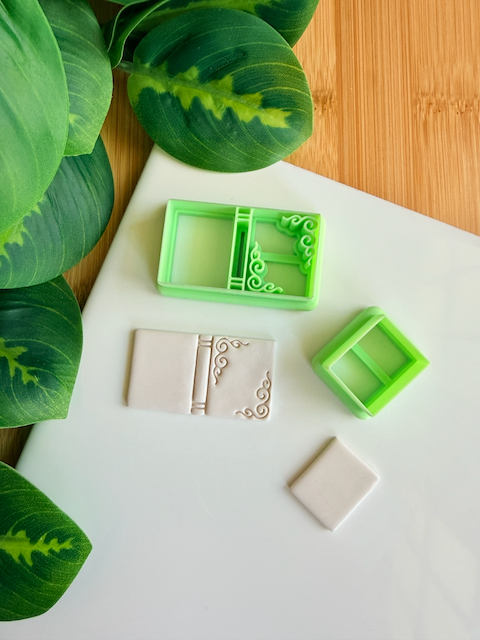 Two light green 3D-printed polymer clay cutters on a white surface. One cutter features an integrated design for a book spine and elegant filigree scrollwork in the corners of the cover. Next to them are two light grey clay samples: one with the embossed ornate book design and a smaller, plain rectangular piece. Green leaves and a wooden surface are visible in the background.