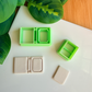Two light green 3D-printed polymer clay cutters on a white surface. One cutter features an integrated design for a book spine and an inset rectangular frame on the cover. Below the tools are two light grey clay samples: one featuring the embossed framed book impression and a smaller, plain rectangular piece. Green leaves and a wooden surface are visible in the background.