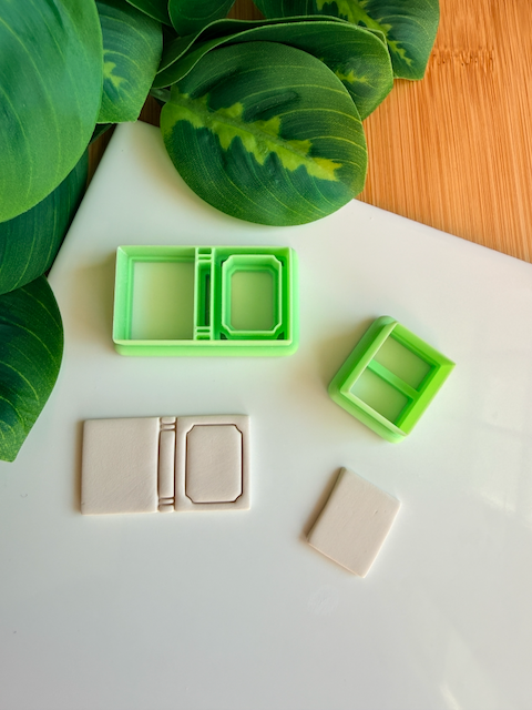Two light green 3D-printed polymer clay cutters on a white surface. One cutter features an integrated design for a book spine and an inset rectangular frame on the cover. Below the tools are two light grey clay samples: one featuring the embossed framed book impression and a smaller, plain rectangular piece. Green leaves and a wooden surface are visible in the background.