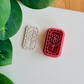 A top-down view of a deep red rectangular clay cutter featuring a central flower-on-a-stem design and a mosaic-tiled border, sitting next to a white polymer clay piece showing the crisp, embossed impression of the same botanical artwork on a clean white surface with vibrant green leaves nearby.