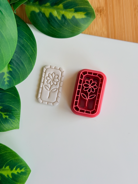 A top-down view of a deep red rectangular clay cutter featuring a central flower-on-a-stem design and a mosaic-tiled border, sitting next to a white polymer clay piece showing the crisp, embossed impression of the same botanical artwork on a clean white surface with vibrant green leaves nearby.