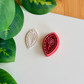 A top-down view of a deep red marquise-shaped clay cutter featuring a central flower-on-a-stem design and a mosaic-tiled border, sitting next to a white polymer clay piece showing the crisp, embossed impression of the same botanical artwork on a clean white surface with vibrant green leaves.