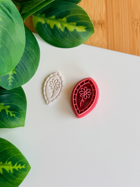 A top-down view of a deep red marquise-shaped clay cutter featuring a central flower-on-a-stem design and a mosaic-tiled border, sitting next to a white polymer clay piece showing the crisp, embossed impression of the same botanical artwork on a clean white surface with vibrant green leaves.