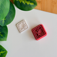 A top-down view of a deep red (or deep reddish-maroon) diamond-shaped clay cutter, featuring a central leafy sprig design and a structured mosaic-tiled border. It is shown sitting next to a matching white polymer clay piece with the corresponding crisp, high-relief embossed botanical impression, arranged on a clean white surface with vibrant green leaves and a light wood surface in the corners.