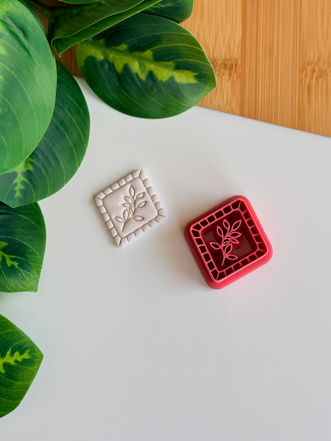 A top-down view of a deep red (or deep reddish-maroon) diamond-shaped clay cutter, featuring a central leafy sprig design and a structured mosaic-tiled border. It is shown sitting next to a matching white polymer clay piece with the corresponding crisp, high-relief embossed botanical impression, arranged on a clean white surface with vibrant green leaves and a light wood surface in the corners.