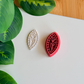 A top-down view of a deep red marquise-shaped clay cutter featuring a central tulip-on-a-stem design and a mosaic-tiled border, shown next to a white polymer clay piece with the corresponding crisp, embossed botanical impression on a clean white surface with vibrant green leaves in the corner.