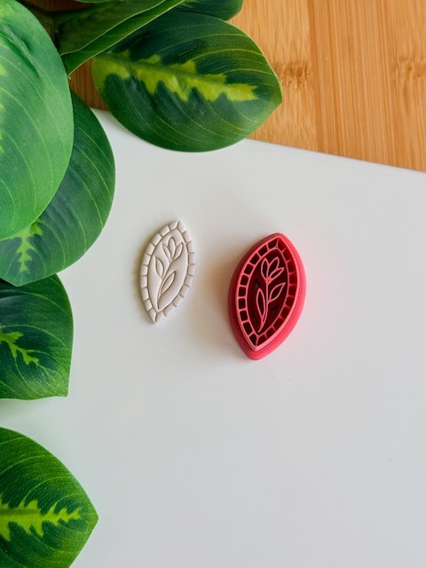A top-down view of a deep red marquise-shaped clay cutter featuring a central tulip-on-a-stem design and a mosaic-tiled border, shown next to a white polymer clay piece with the corresponding crisp, embossed botanical impression on a clean white surface with vibrant green leaves in the corner.