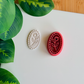 A magenta 3D-printed oval clay cutter and debosser featuring a single rose design inside an ornate beaded frame. To the left, a light grey clay sample shows the crisp, embossed floral impression and the textured border. The items are displayed on a white surface with green leaves and a wooden background visible in the corners.