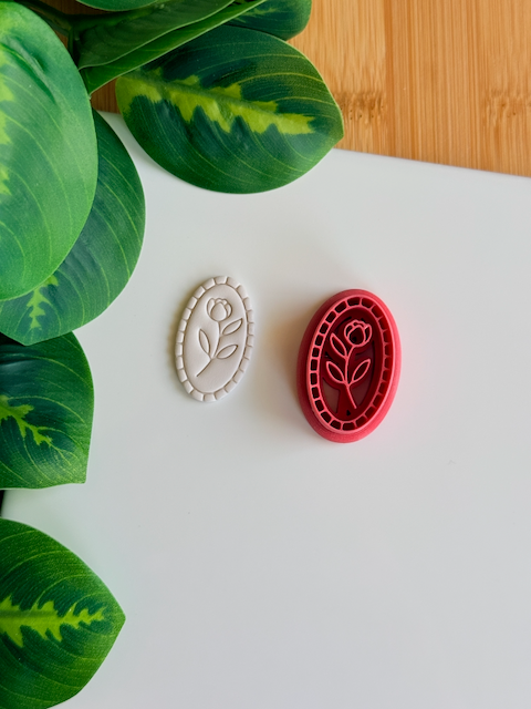 A magenta 3D-printed oval clay cutter and debosser featuring a single rose design inside an ornate beaded frame. To the left, a light grey clay sample shows the crisp, embossed floral impression and the textured border. The items are displayed on a white surface with green leaves and a wooden background visible in the corners.
