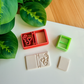 A reddish-orange 3D-printed polymer clay cutter with an integrated design for a book spine and an elegant heart scroll motif on the cover. Below it is a light grey clay sample with the embossed romantic book design. A small light green rectangular cutter and a plain rectangular clay piece are also shown on the white surface, with green leaves and a wooden surface in the background.