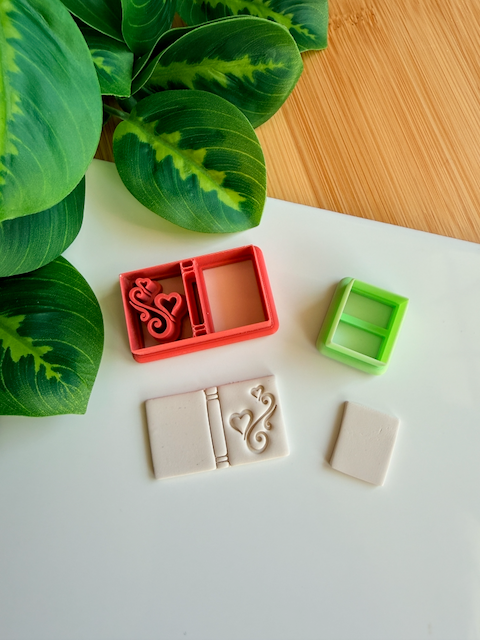 A reddish-orange 3D-printed polymer clay cutter with an integrated design for a book spine and an elegant heart scroll motif on the cover. Below it is a light grey clay sample with the embossed romantic book design. A small light green rectangular cutter and a plain rectangular clay piece are also shown on the white surface, with green leaves and a wooden surface in the background.