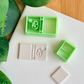 Two light green 3D-printed polymer clay cutters on a white surface. One cutter includes an integrated stamp of a book spine and a lock-and-key motif. Next to the tools are two samples of light grey clay: one featuring the detailed book impression and one plain rectangular piece. Green leaves are visible in the corner of the frame.