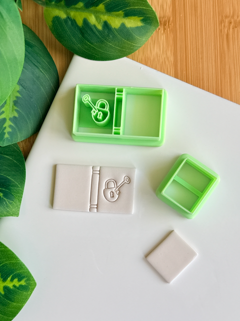 Two light green 3D-printed polymer clay cutters on a white surface. One cutter includes an integrated stamp of a book spine and a lock-and-key motif. Next to the tools are two samples of light grey clay: one featuring the detailed book impression and one plain rectangular piece. Green leaves are visible in the corner of the frame.