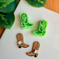 A mirrored pair of light green 3D-printed cowboy boot clay cutters with an intricate leaf and vine "embroidery" pattern. Below the tools are two finished brown clay boot samples featuring the embossed design and decorated with small handmade white flowers at the ankle. The scene is framed by green leaves on a white surface with a wooden background.