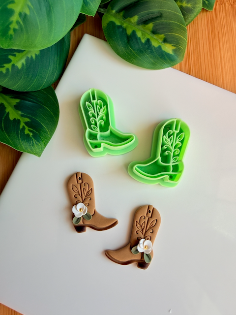 A mirrored pair of light green 3D-printed cowboy boot clay cutters with an intricate leaf and vine "embroidery" pattern. Below the tools are two finished brown clay boot samples featuring the embossed design and decorated with small handmade white flowers at the ankle. The scene is framed by green leaves on a white surface with a wooden background.