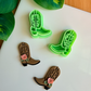 A mirrored pair of light green 3D-printed cowboy boot clay cutters with an intricate leaf and vine "embroidery" pattern. Below the tools are two finished brown clay boot samples featuring the embossed design and decorated with small handmade white flowers at the ankle. The scene is framed by green leaves on a white surface with a wooden background.