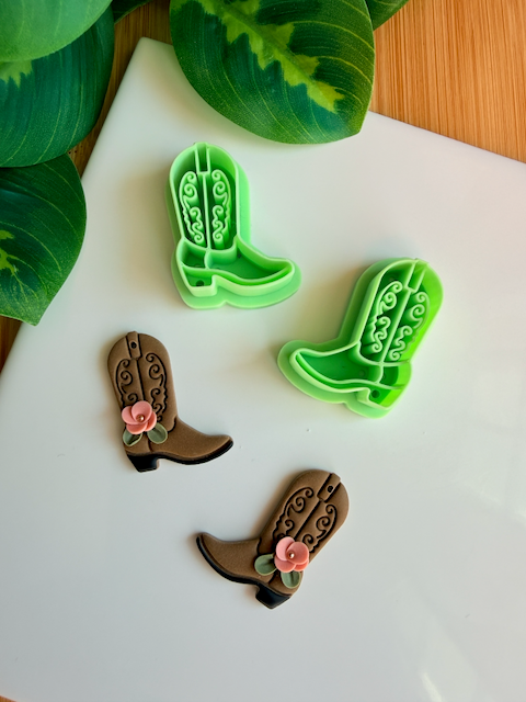 A mirrored pair of light green 3D-printed cowboy boot clay cutters with an intricate leaf and vine "embroidery" pattern. Below the tools are two finished brown clay boot samples featuring the embossed design and decorated with small handmade white flowers at the ankle. The scene is framed by green leaves on a white surface with a wooden background.