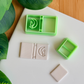A top-down view of a mint green, multi-piece 3D-printed clay cutter set featuring a "moonlit journal" theme. The main cutter creates an open book shape with a central banded spine and an emblem of a cat sitting on a crescent moon, shown alongside a white polymer clay piece featuring the crisp, embossed impression of the same celestial artwork. A smaller rectangular cutter and a plain clay piece are also included in the arrangement on a clean white surface with green leaves and a wooden background in the cor