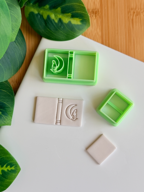 A top-down view of a mint green, multi-piece 3D-printed clay cutter set featuring a "moonlit journal" theme. The main cutter creates an open book shape with a central banded spine and an emblem of a cat sitting on a crescent moon, shown alongside a white polymer clay piece featuring the crisp, embossed impression of the same celestial artwork. A smaller rectangular cutter and a plain clay piece are also included in the arrangement on a clean white surface with green leaves and a wooden background in the cor