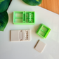 Two light green 3D-printed polymer clay cutters on a white surface. One cutter features an integrated design for a book spine and a vertical row of moon phases on the cover. Next to the tools are two light grey clay samples: one with the embossed celestial book design and a smaller, plain rectangular piece. Green leaves and a wooden surface are visible in the background.