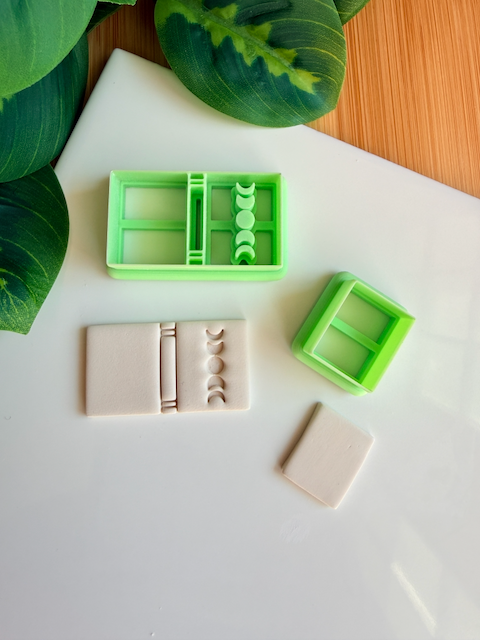 Two light green 3D-printed polymer clay cutters on a white surface. One cutter features an integrated design for a book spine and a vertical row of moon phases on the cover. Next to the tools are two light grey clay samples: one with the embossed celestial book design and a smaller, plain rectangular piece. Green leaves and a wooden surface are visible in the background.